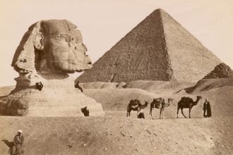 grayscale photo of pyramid and camels in the desert
